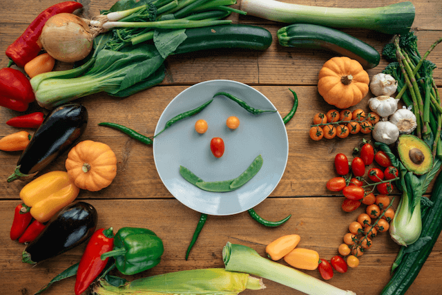 Vegetables placed on a plate representing a human face with arms and legs surrounded by other vegetables in a circle.