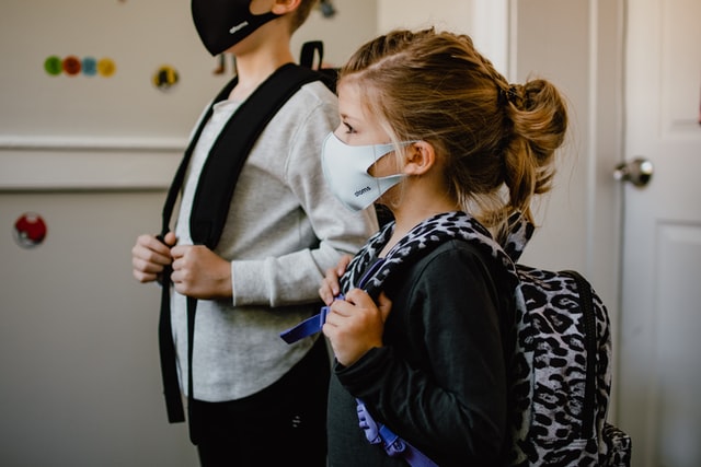 A small boy and a girl wearing face masks with bags on their shoulders.