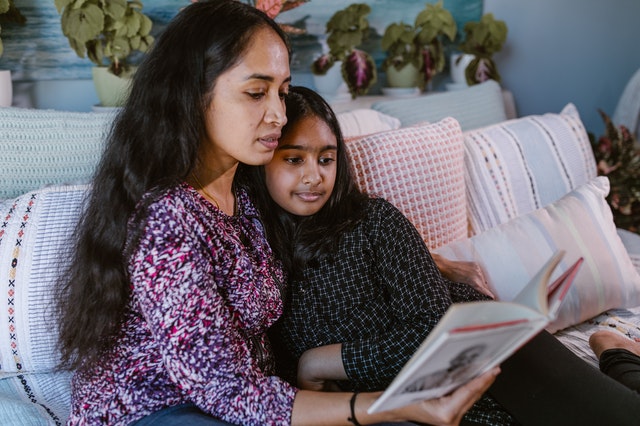 Indian mother and daughter reading a book while sitting on bed.
